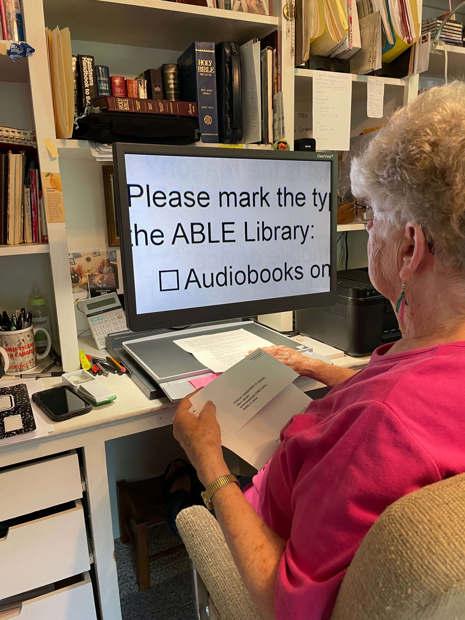 An older woman in a pink shirt sits at a desk, reading a paper with the help of a large screen magnifier displaying text about audiobooks from the ABLE Library. The workspace is filled with books, papers, and office supplies.