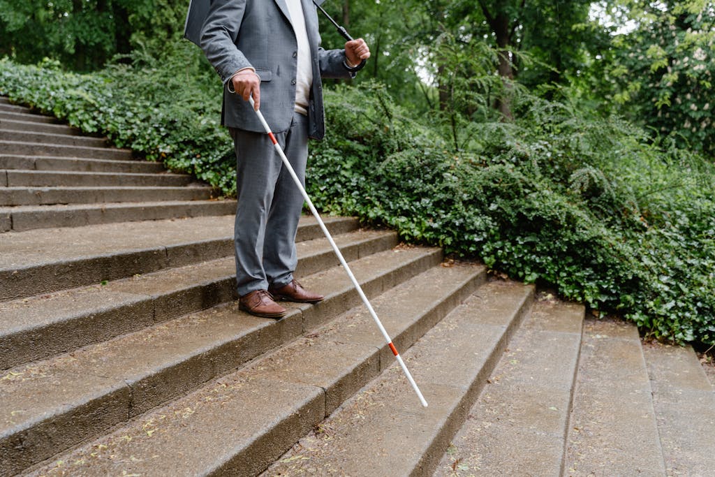 A man uses a white cane to navigate concrete stairs in a lush green setting, symbolizing independence.
