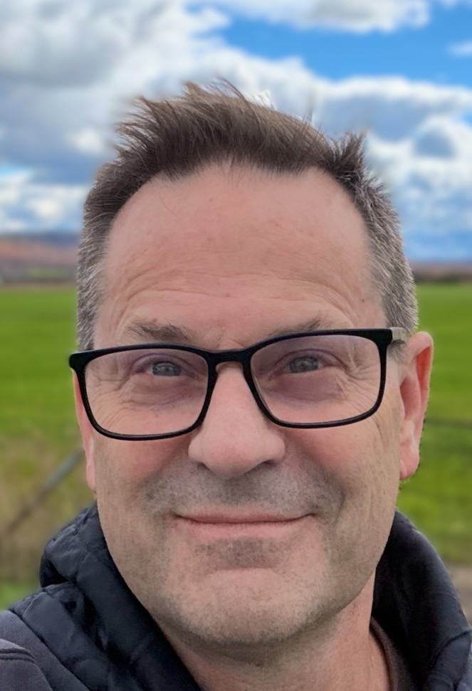A middle-aged man with short brown hair and black glasses smiles outdoors in front of a green field and a partly cloudy sky.