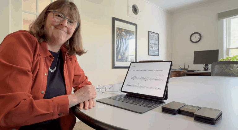 A woman sits at a round table in an office, smiling at the camera. In front of her is a tablet with sheet music on the screen and several small black devices. The background shows a desk, clock, and framed pictures on the wall.