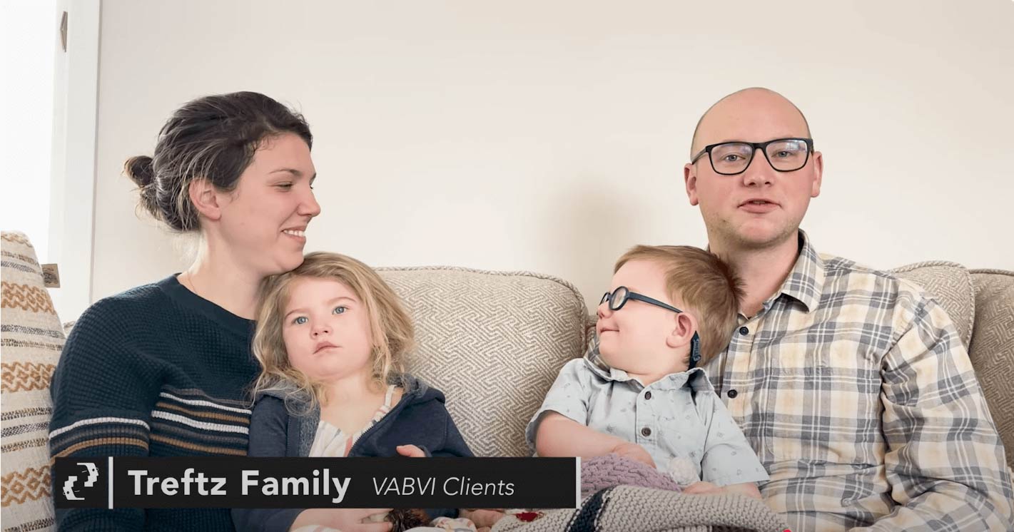 A family of four sits on a couch. The mother holds a young girl, while the father, wearing glasses, sits beside a young boy who is also wearing glasses. Text reads: Trefz Family VABVI Clients.
