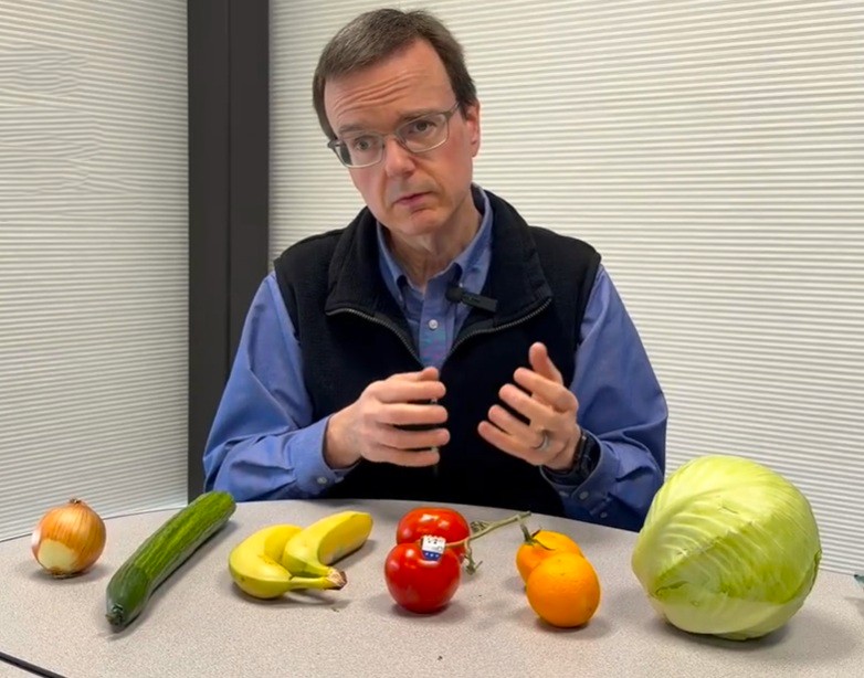 A man with glasses sits at a table with an onion, cucumber, bananas, two tomatoes, an orange, and a cabbage arranged in front of him. He is gesturing with his hands while looking at the camera.