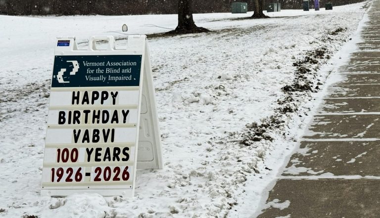A sandwich board sign on a snowy sidewalk reads: Vermont Association for the Blind and Visually Impaired. Happy Birthday VABVI. 100 Years. 1926-2026. Snow covers the ground and trees line the background.