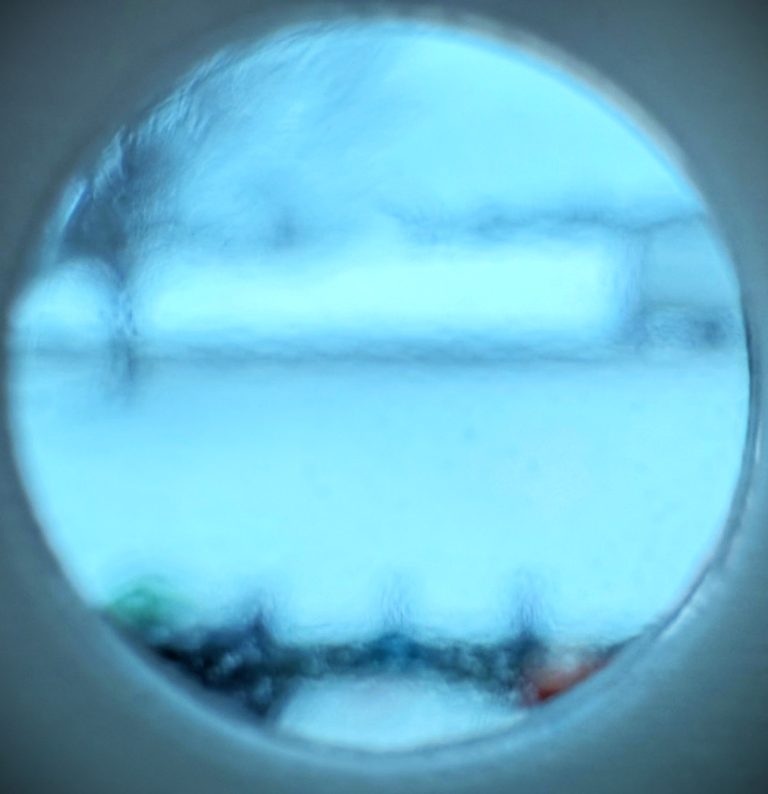 A blurry, circular view through frosted glass shows a wintry, snow-covered landscape with indistinct objects and trees, all tinted in cool blue tones.