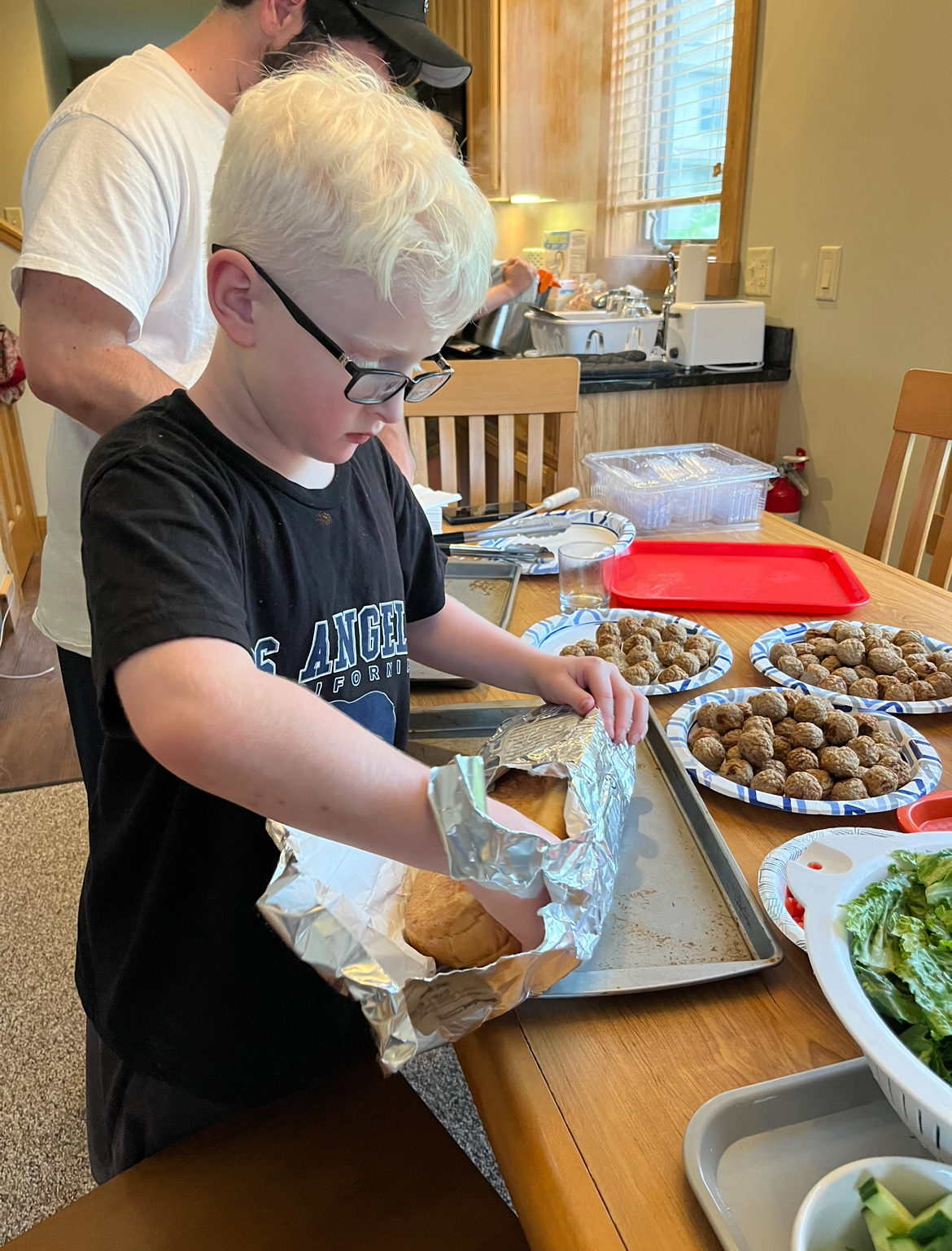 A young boy with blond hair and glasses unwraps foil from a loaf of bread at a table set with food, including salad and plates of falafel. An adult stands nearby in a kitchen setting.