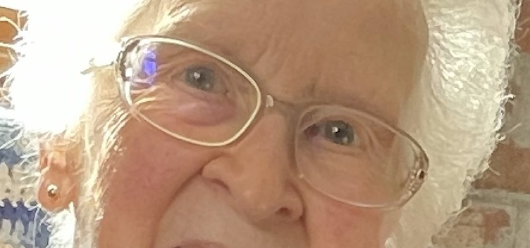 Close-up of an elderly woman with white hair, wearing glasses and a small earring, smiling gently. A crochet blanket and brick wall are visible in the background.
